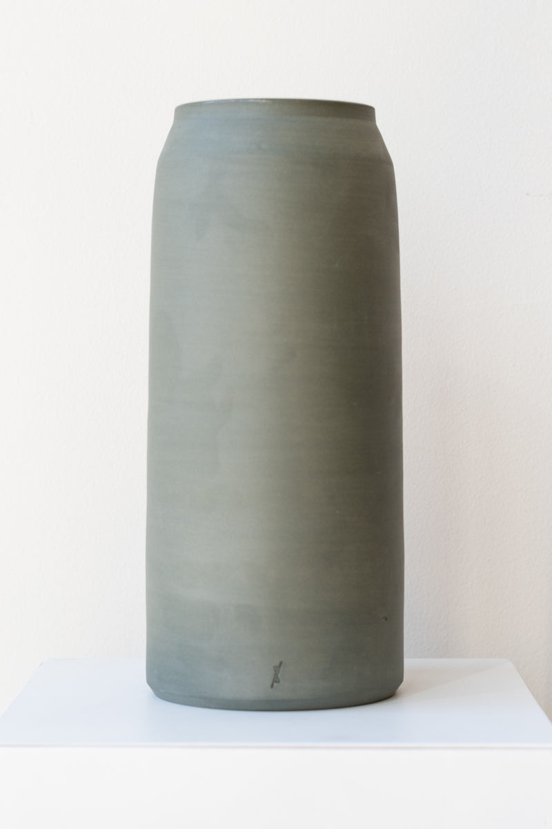 One green-grey stoneware vase sits on a white surface in a white room. It is round and tall, and it has a small logo imprinted at the base of the vase. It is photographed straight on.