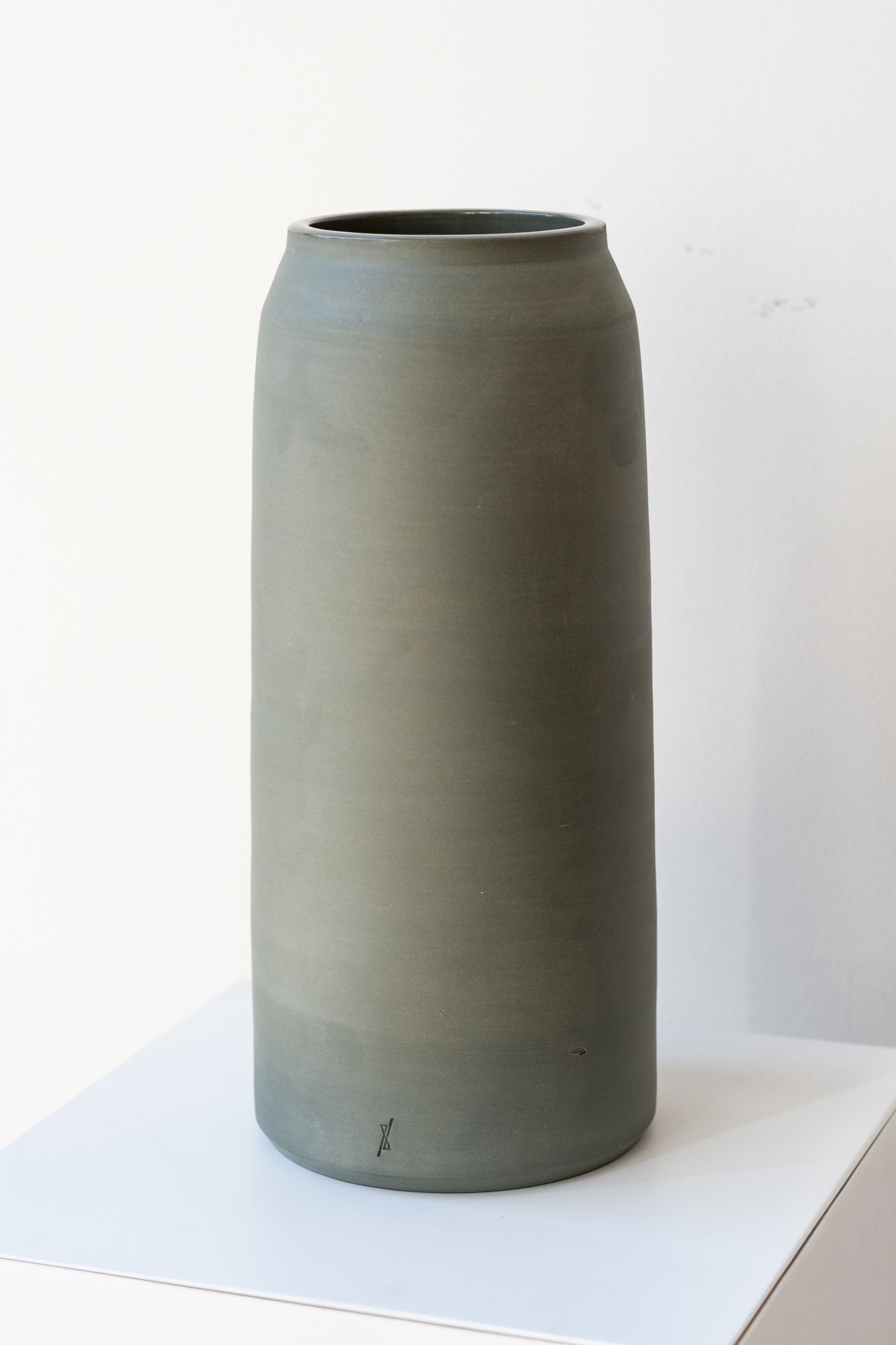 One green-grey stoneware vase sits on a white surface in a white room. It is round and tall, and it has a small logo imprinted at the base of the vase. It is photographed at a slight angle to show the detail in the clay.