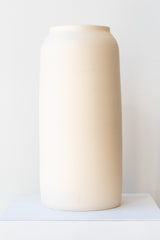 One cream colored bouquet vase sits on a white surface in a white room. The vase is tall and cylindrical, with a slight rim at the top. It is photographed straight on.