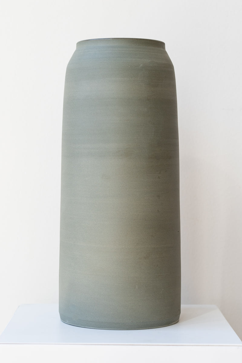 One green-grey stoneware vase sits on a white surface in a white room. It is round and tall, and it has a small logo imprinted at the base of the vase. It is photographed straight on.