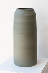 One green-grey stoneware vase sits on a white surface in a white room. It is round and tall, and it has a small logo imprinted at the base of the vase. It is photographed at a slight angle to show the detail in the clay.
