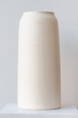 One large cream colored bouquet vase sits on a white surface in a white room. The vase is tall and cylindrical, with a slight rim at the top. It is photographed straight on.