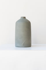 One grey stoneware bud vase sits on a white surface in a white room. It is short and cylindrical with a narrow opening at the top. It has a tiny logo imprinted in the bottom of the clay. It is photographed straight on.