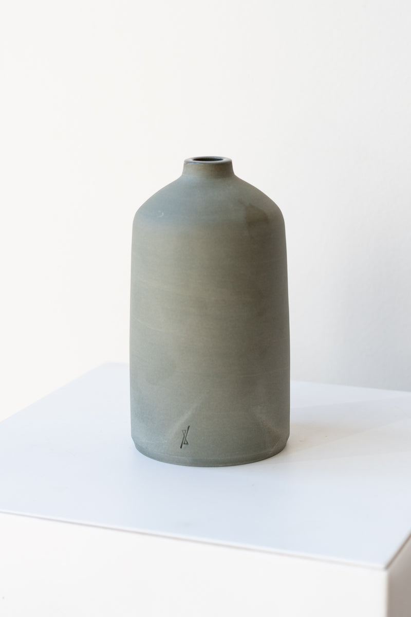 One grey stoneware bud vase sits on a white surface in a white room. It is short and cylindrical with a narrow opening at the top. It has a tiny logo imprinted in the bottom of the clay. It is photographed at a slight angle to show details in the clay.