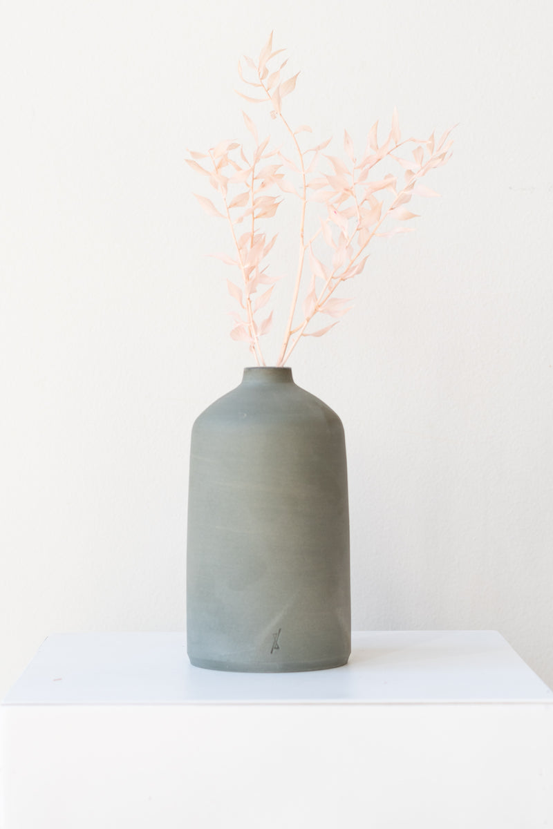 One grey stoneware bud vase sits on a white surface in a white room. It is short and cylindrical with a narrow opening at the top. It has a tiny logo imprinted in the bottom of the clay. The bud vase has a small sprig of light pink foliage in it. It is photographed straight on.