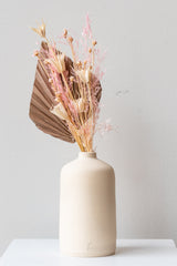 One white stoneware bud vase sits on a white surface in a white room. It is short and cylindrical with a narrow opening at the top. It has a tiny logo imprinted in the bottom of the clay. Inside the vase is a tan, cream, and pink dried floral arrangement. It is photographed straight on.