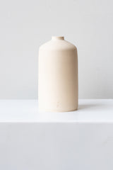 One white stoneware bud vase sits on a white surface in a white room. It is short and cylindrical with a narrow opening at the top. It has a tiny logo imprinted in the bottom of the clay. It is photographed straight on.