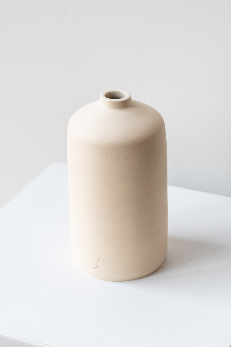 One white stoneware bud vase sits on a white surface in a white room. It is short and cylindrical with a narrow opening at the top. It has a tiny logo imprinted in the bottom of the clay. It is photographed closer and at an angle.