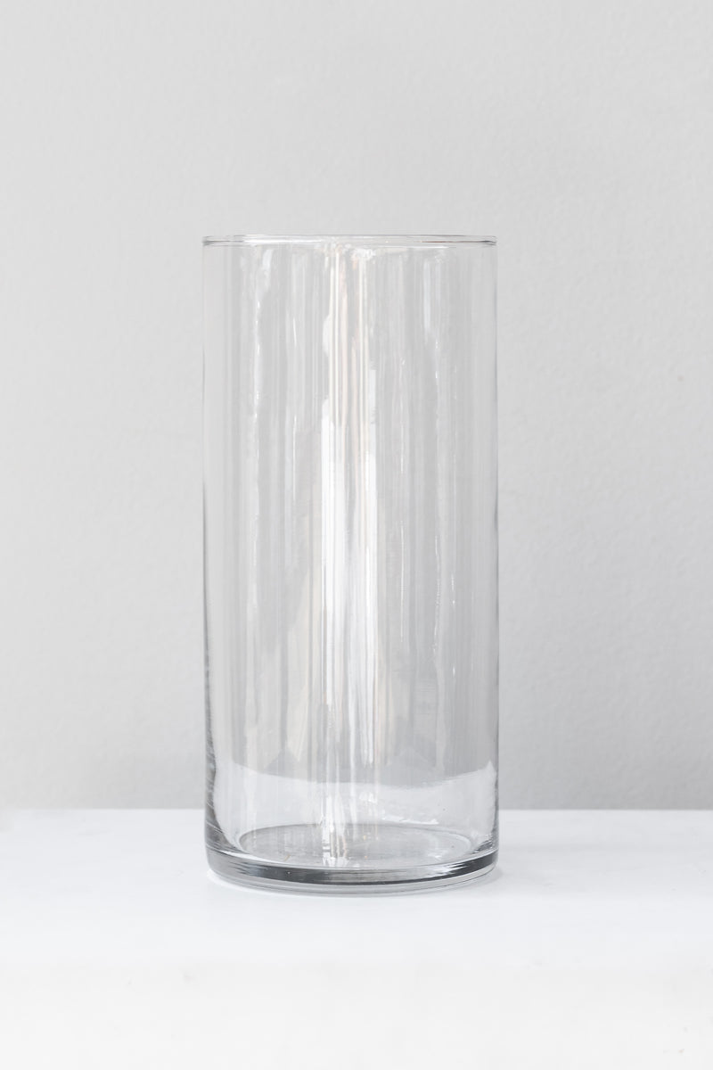 Clear cylindrical glass vase sits on a white surface in a white room. It is photographed straight on.