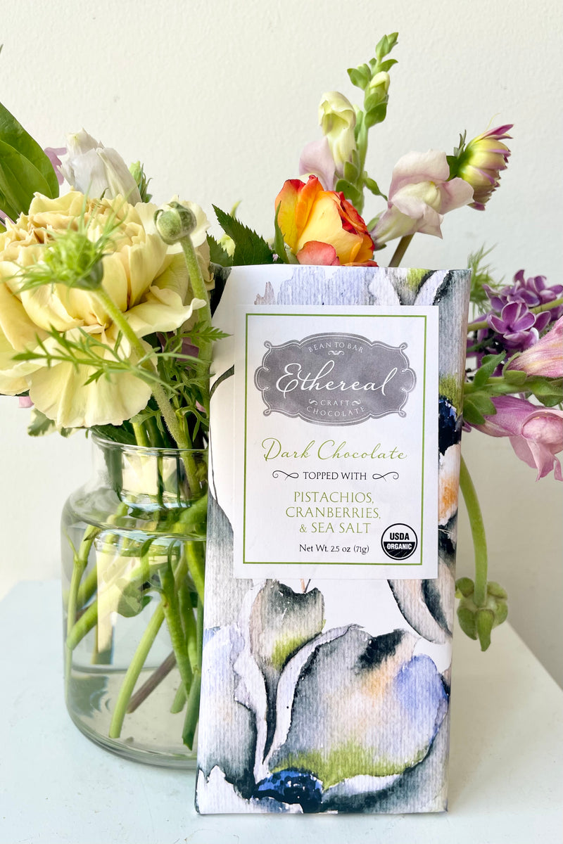 Ethereal Chocolate dark chocolate bar with pistachios, cranberries, and sea salt behind flowers on a white surface in a white room