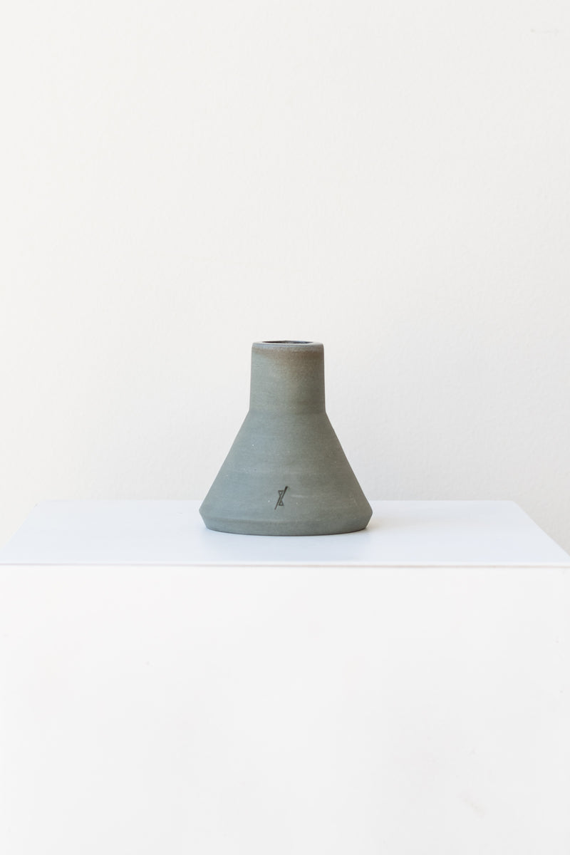 One triangular grey stoneware candle holder sits on a white, flat surface in a white room. It has a small logo etched into the clay near the base. It is photographed straight on. 