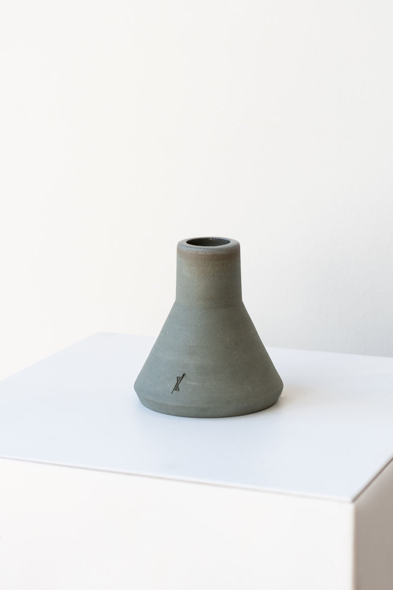 One triangular grey stoneware candle holder sits on a white, flat surface in a white room. It has a small logo etched into the clay near the bottom. It is photographed from a slight angle.