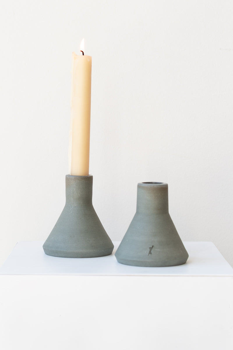 Two triangular grey stoneware candle holders sit on a white surface in a white room. The candle holder on the left has an ivory taper candle, which is lit. The candle holder on the right has a small logo etched into the clay near the bottom. They are photographed straight on.