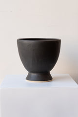 One glazed stoneware planter sits on a white surface in a white room. The planter is matte black. The planter has a small angled base with a larger bowl shaped top. It is photographed straight on.