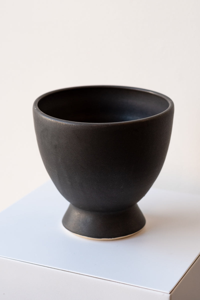 One glazed stoneware planter sits on a white surface in a white room. The planter is matte black. The planter has a small angled base with a larger bowl shaped top. It is photographed closer and at a slight angle.