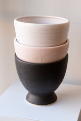Three glazed stoneware planters are stacked on a white surface in a white room. The bottom planter is matte black, the second planter is light pink, and the top planter is white with tiny black specks. They are photographed at a slight angle to show the texture in the glaze.