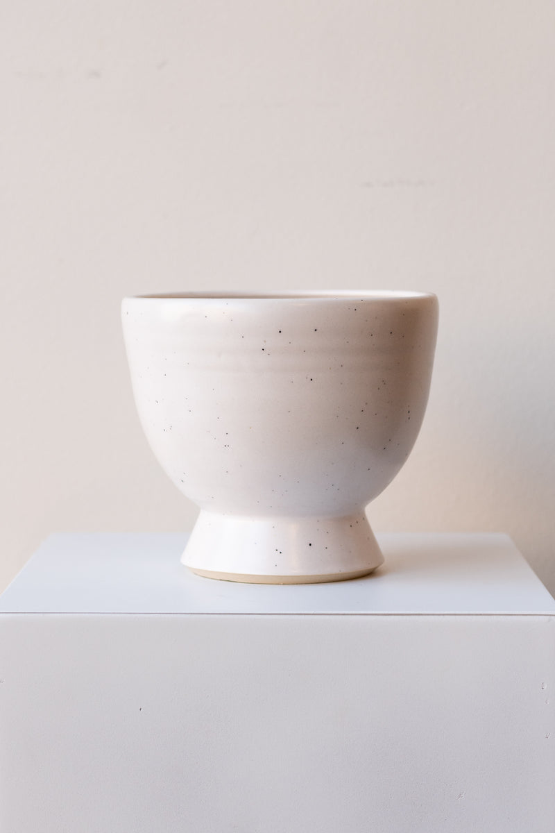 One glazed stoneware planter sits on a white surface in a white room. The planter is white with tiny black specks. The planter has a small angled base with a larger bowl shaped top. It is photographed straight on.