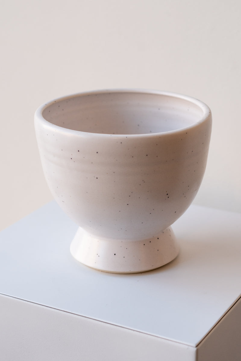 One glazed stoneware planter sits on a white surface in a white room. The planter is white with tiny black specks. The planter has a small angled base with a larger bowl shaped top. It is photographed closer and at a slight angle.