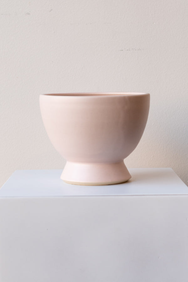 One glazed stoneware planter sits on a white surface in a white room. The planter is light pink. The planter has a small angled base with a larger bowl shaped top. It is photographed straight on.