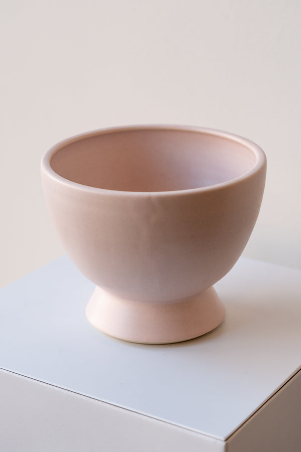 One glazed stoneware planter sits on a white surface in a white room. The planter is light pink. The planter has a small angled base with a larger bowl shaped top. It is photographed closer and at a slight angle.