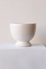 One glazed stoneware planter sits on a white surface in a white room. The planter is white. The planter has a small angled base with a larger bowl shaped top. It is photographed straight on.