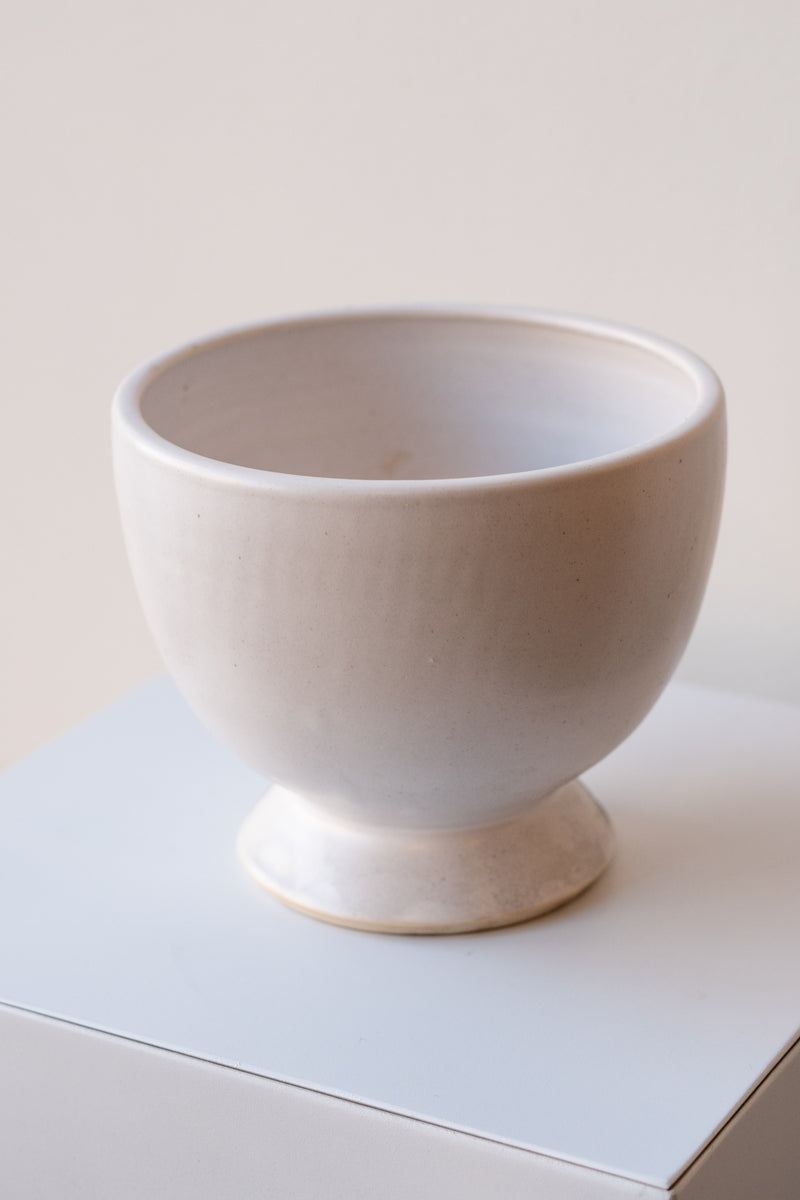 One glazed stoneware planter sits on a white surface in a white room. The planter is white. The planter has a small angled base with a larger bowl shaped top. It is photographed closer and at a slight angle.