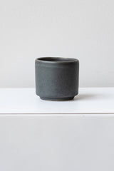 A small grey cup planter sits on a white surface in a white room. It is photographed straight on.