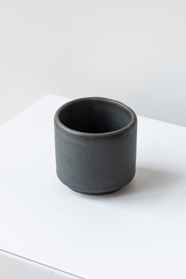 A small grey cup planter sits on a white surface in a white room. It is photographed closer and at an angle.