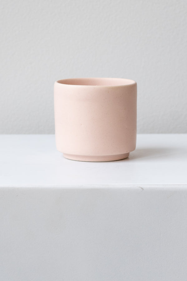 A small pink cup planter sits on a white surface in a white room. It is photographed straight on.