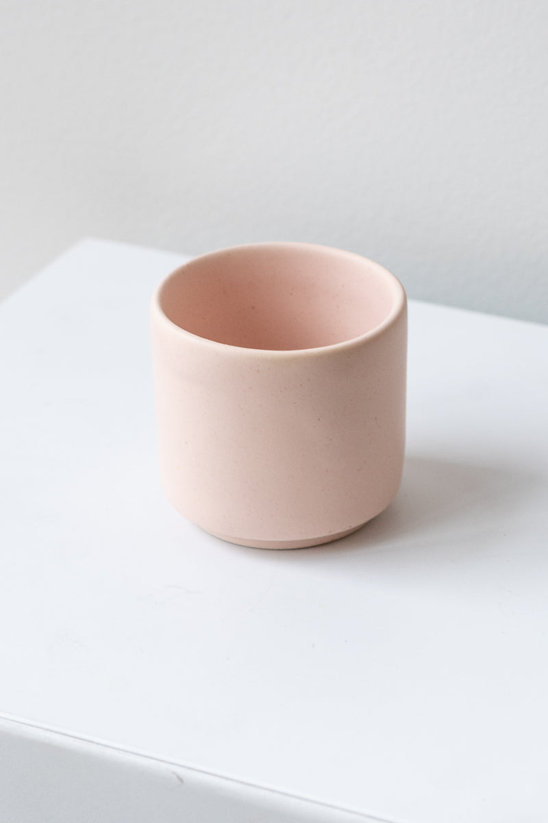 A small pink cup planter sits on a white surface in a white room. It is photographed closer and at an angle.
