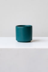 A small dark teal cup planter sits on a white surface in a white room. It is photographed straight on.