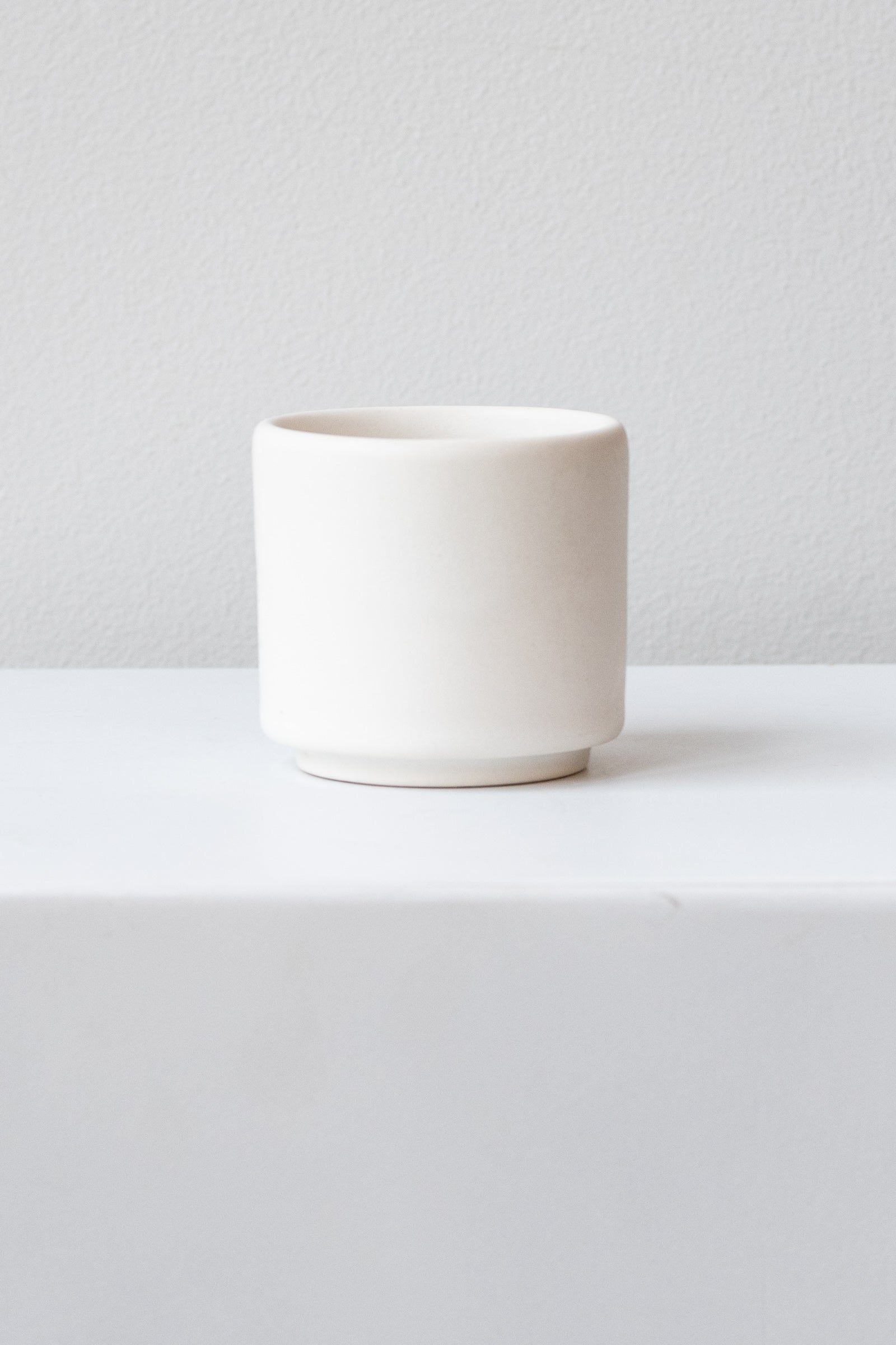 A small white cup planter sits on a white surface in a white room. It is photographed straight on.