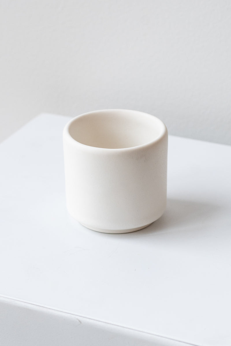 A small white cup planter sits on a white surface in a white room. It is photographed closer and at an angle.