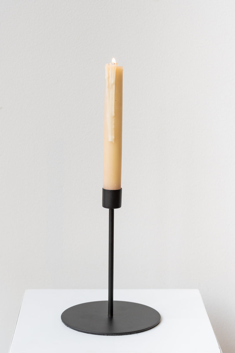 Black tall taper candle holder on a white surface in a white room - inside the candle holder is a lit beige taper candle