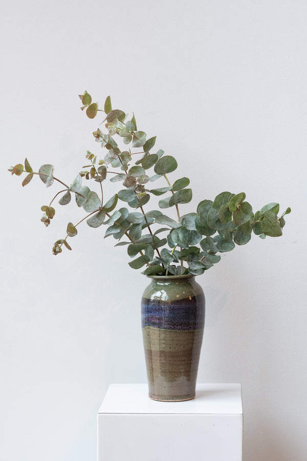Bruning Pottery moss and lilac glazed tall ceramic vase in front of white background. Inside the vase is a bunch of fresh eucalyptus