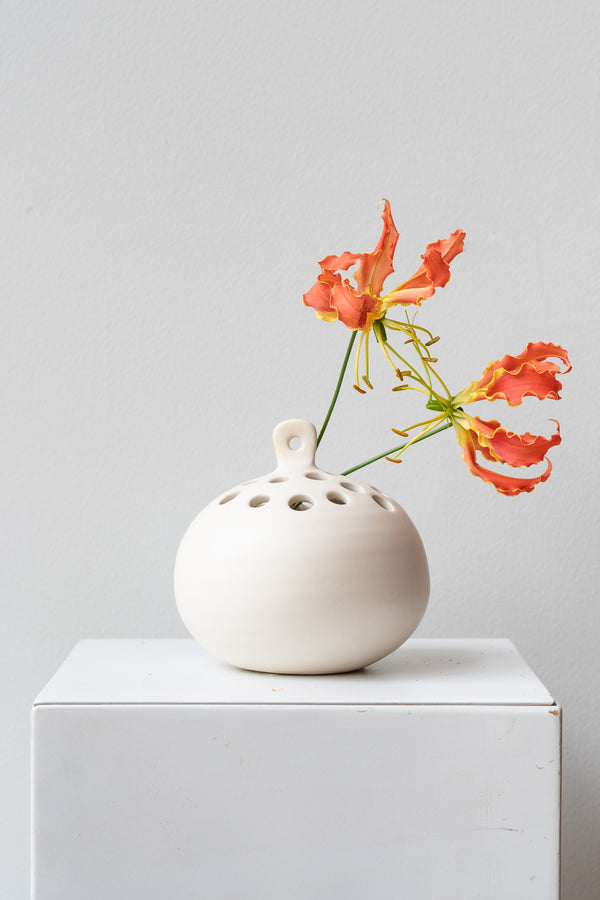 White Ceramic Hanging Flower Frog Vase on a white surface in a white room. Inside the vase are two orange Gloriosa Lillies
