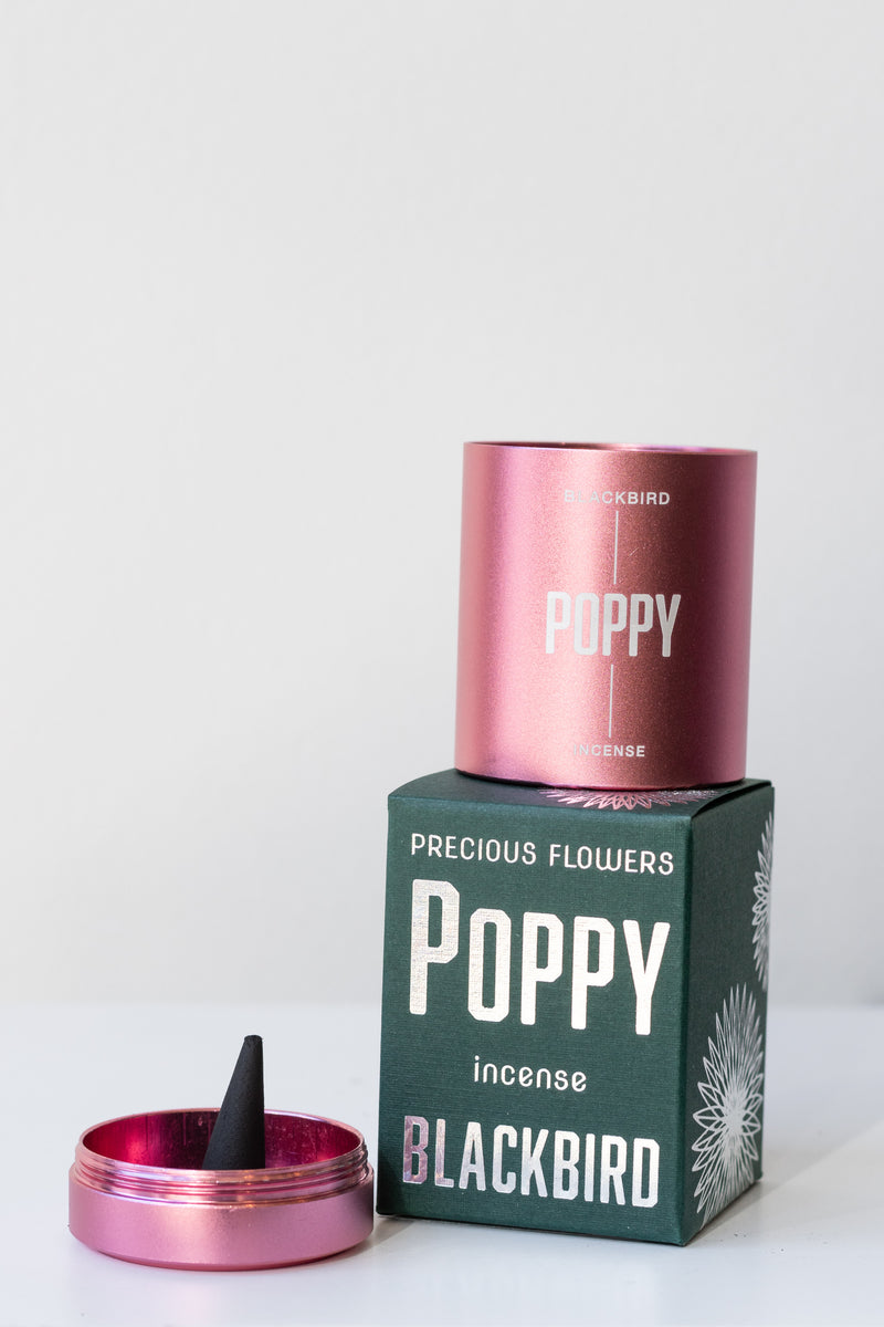 Box of Poppy incense by Blackbird in front of white background. On top of the box is the pink metal cylindrical incense tin without its lid, and to the left of the box is the turned-over lid acting as an incense burner with a single incense cone inside.