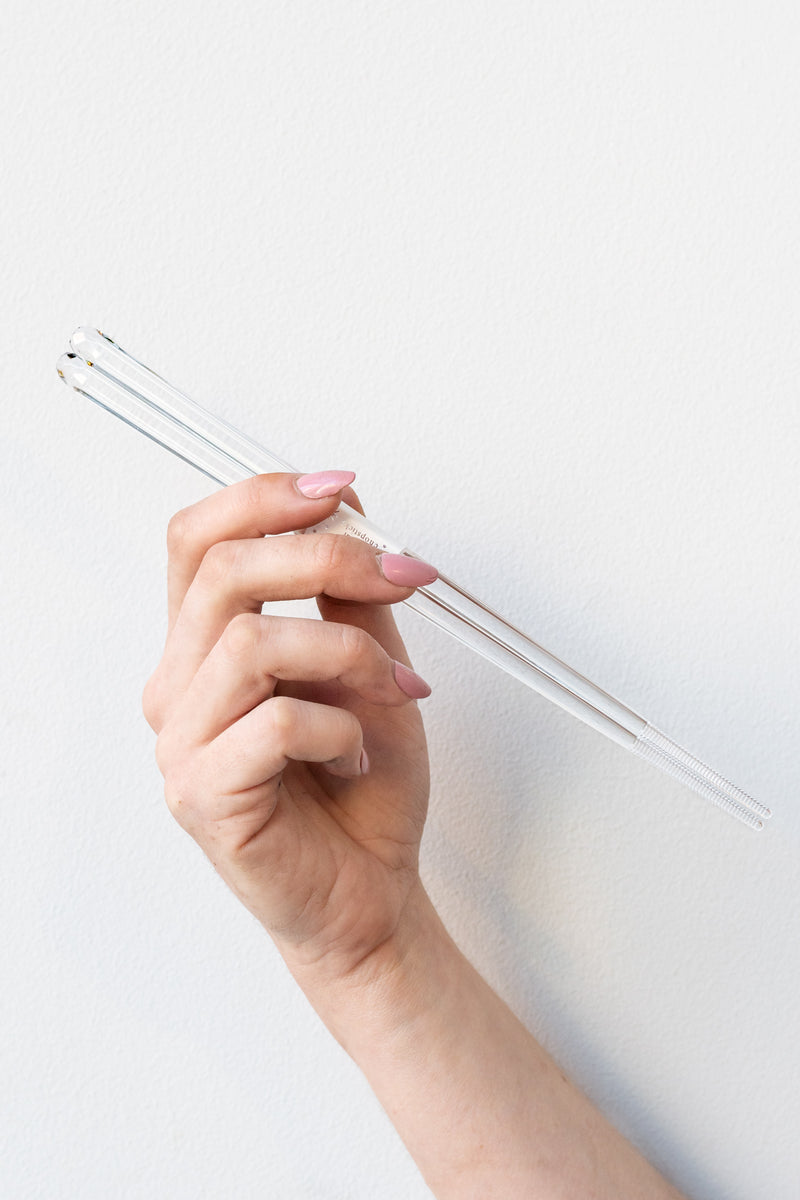 White hand with pink nail polish holding a set of clear acrylic chopsticks in front of a white background