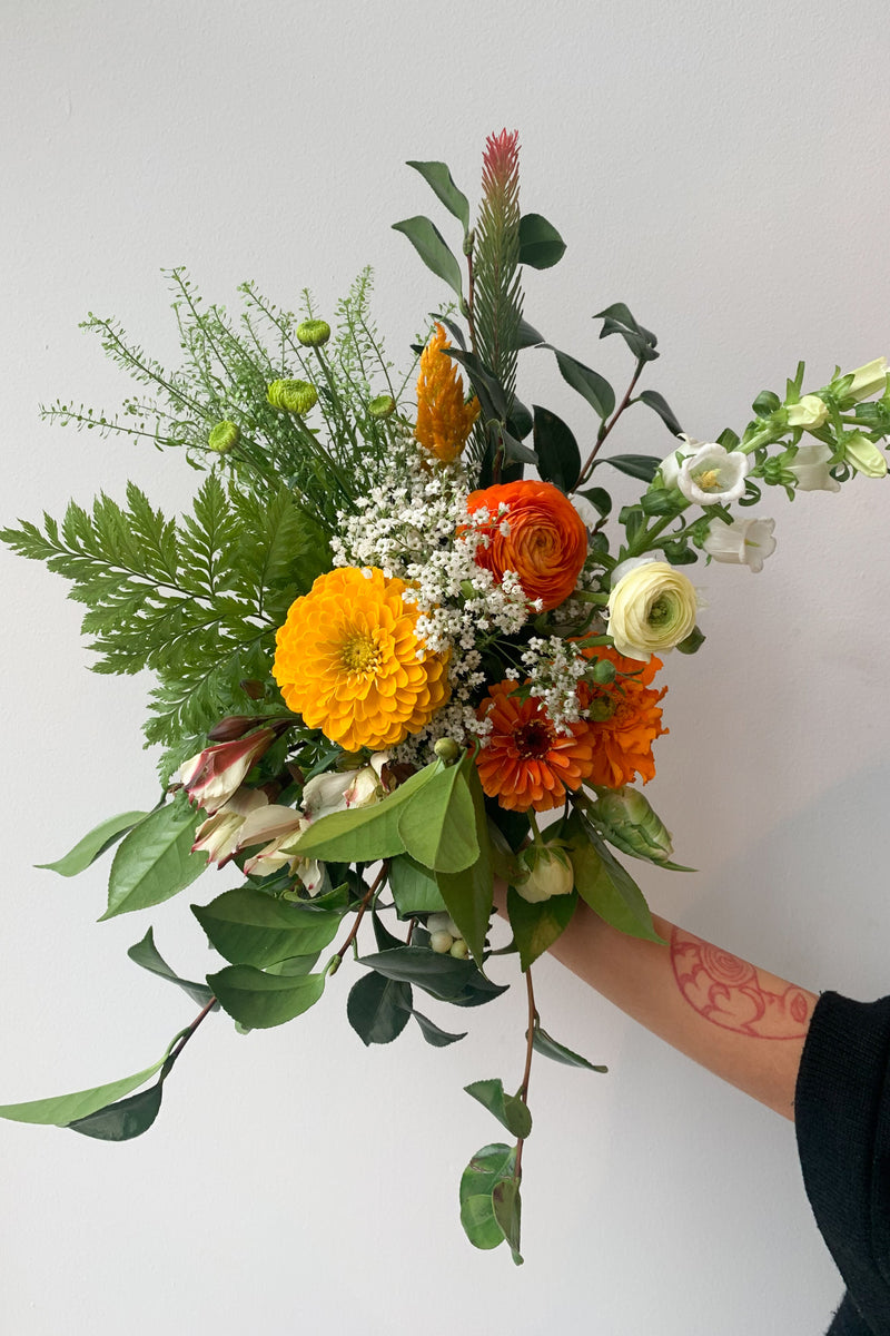 Colorful orange, yellow and white floral sprays and greenery being held by hand in a custom floral arrangement by Sprout Home. 