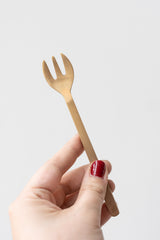 Fog Linen Work brass dessert fork held by white person's hand with red nails in front of white background