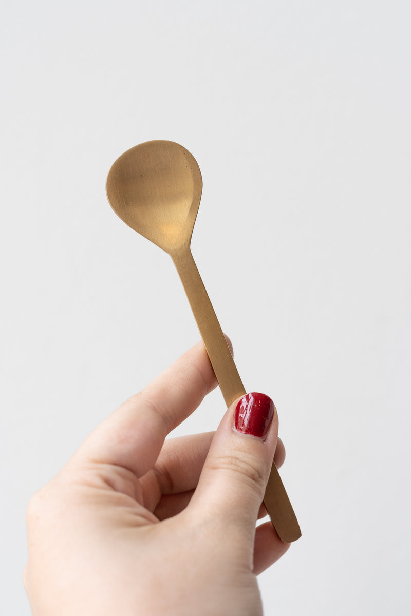 Fog Linen Work brass dessert spoon held by white person's hand with red nails in front of white background