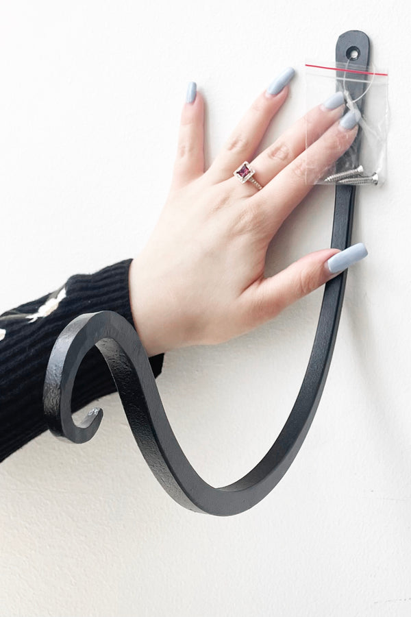A hand holds Upcurled Metal Wallhook 12" against white wall