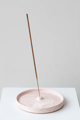 The Northern Habitat Incense Tray blush marble on a white surface in a white room. Inside the holder is a single stick of incense