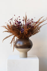 OYOY Living Design Hagi brown vase on white surface in a white room. Inside the vase is a brown, red, and purple dried floral arrangement