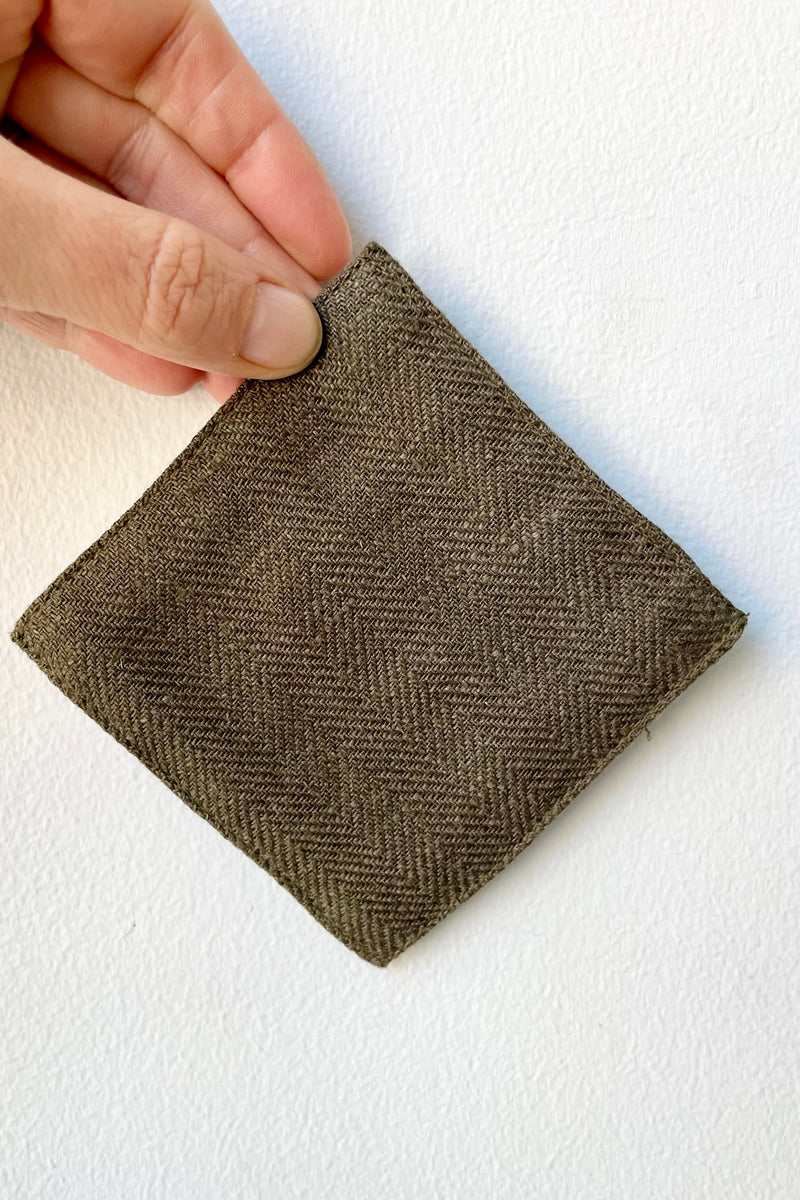 A hand holds the Herringbone Green Linen Coaster against a white backdrop.