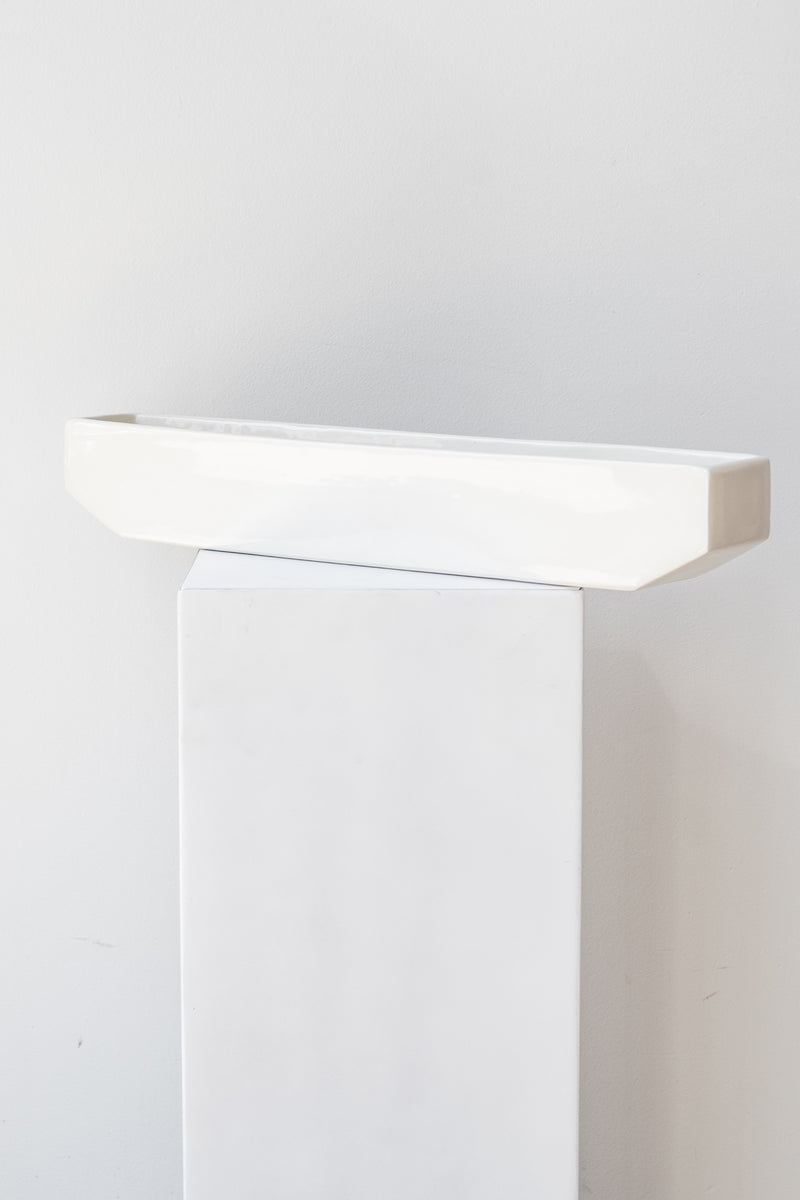 One long rectangular white planter sits on a white square pedestal in a white room. The planter is empty. It is photographed straight on.