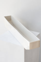 One long rectangular white planter sits on a white square pedestal in a white room. The planter is empty. It is photographed closer and at an angle.