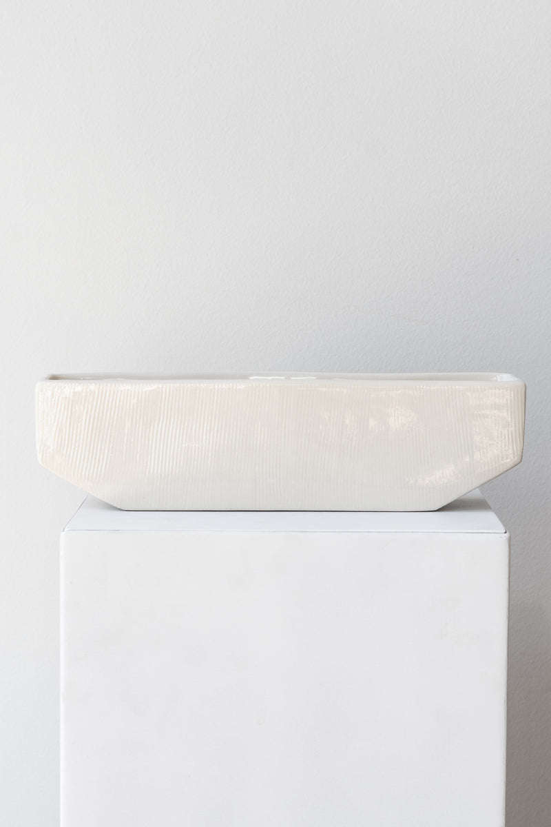 One medium rectangular white planter sits on a white square pedestal in a white room. The planter is empty. It is photographed straight on.