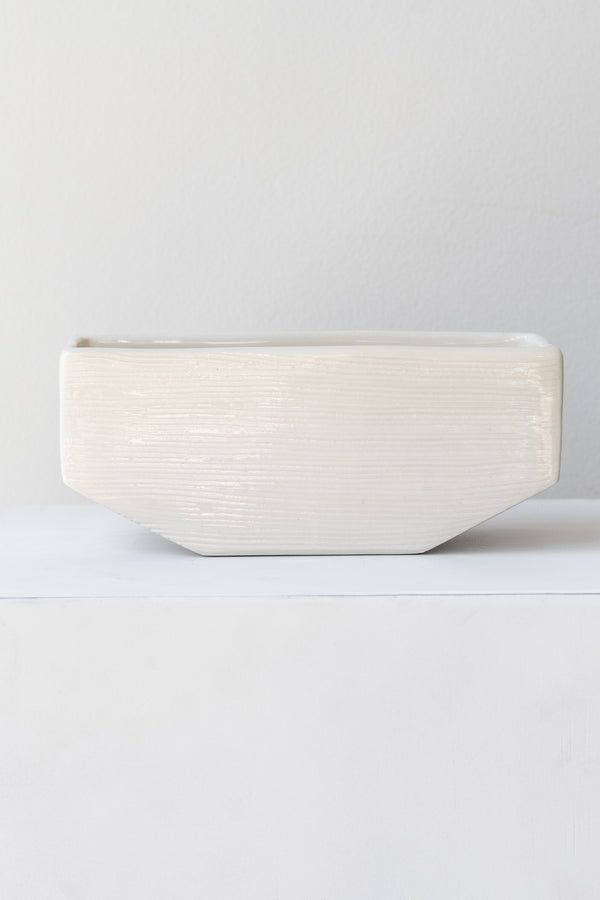 One small rectangular white planter sits on a white square pedestal in a white room. The planter is empty. It is photographed straight on.
