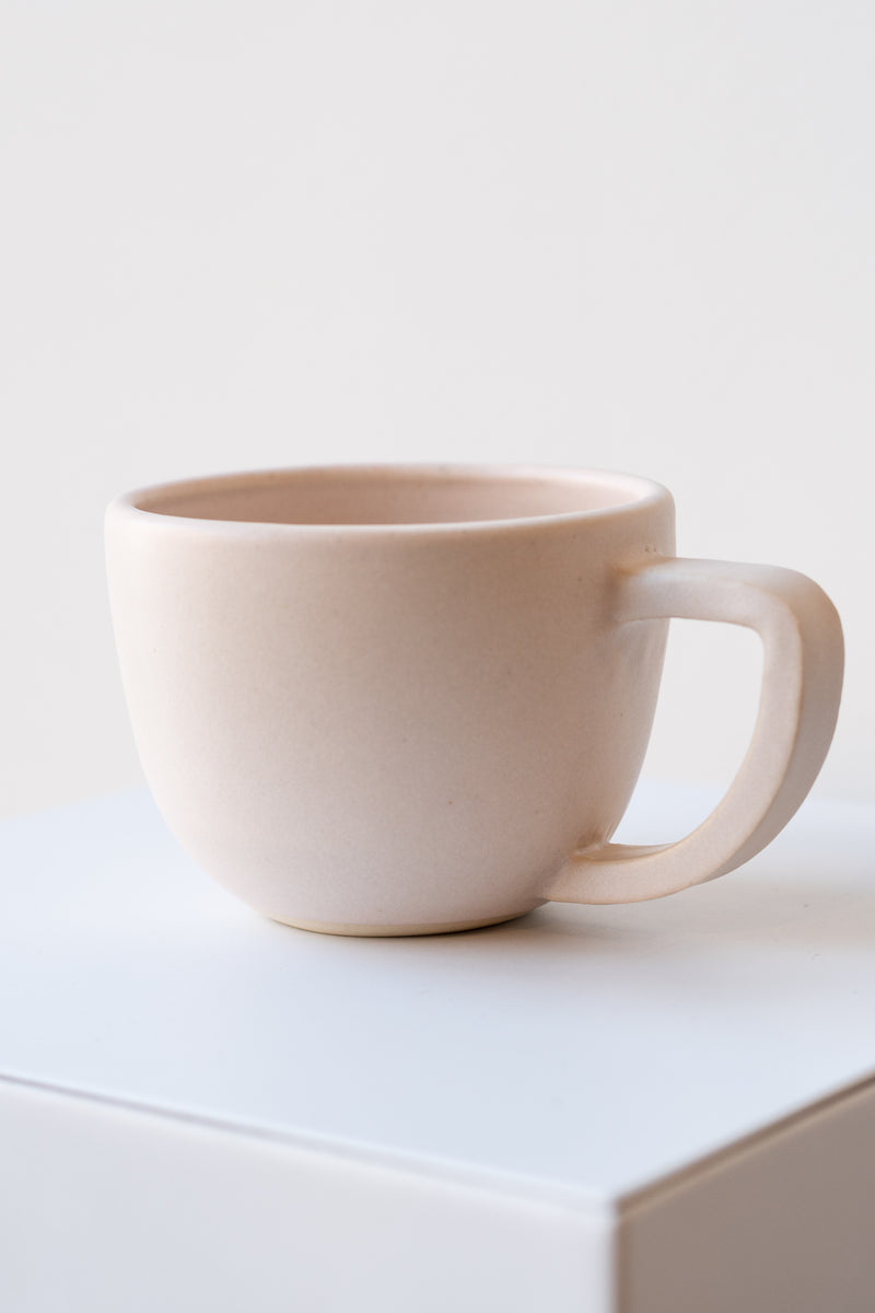One ceramic mug sits on a white surface in a white room. The mug is light pink. The mug is empty. Is is photographed closer and at an angle to show the shape of the handle, which is rounded with a flat top.